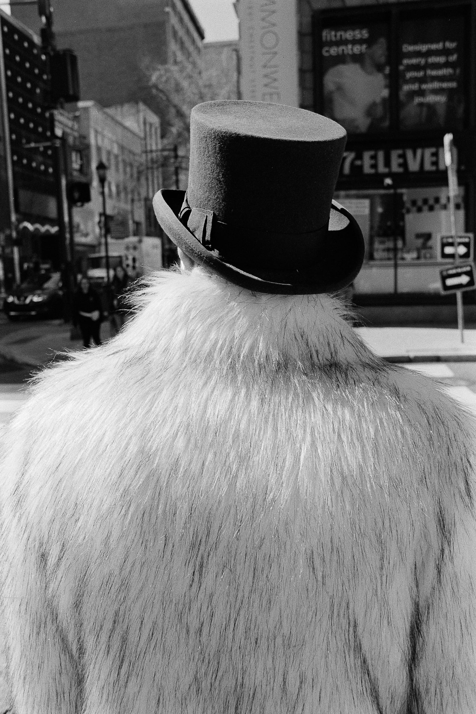A person seen from behind in a tall black top hat and a shaggy white coat at a street corner outside a 7-Eleven