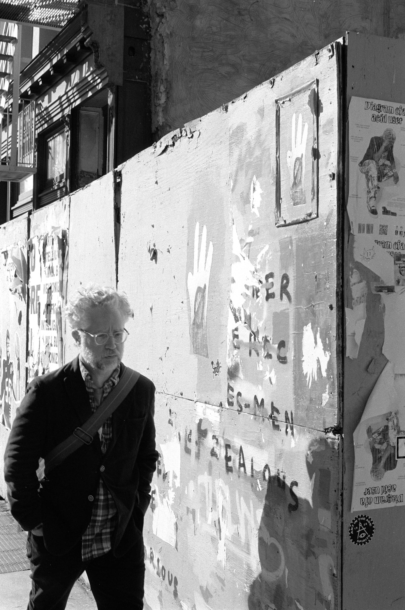 A grey-haired man with a shoulder bag walking past a plywood hoarding covered in torn flyposters and painted hands