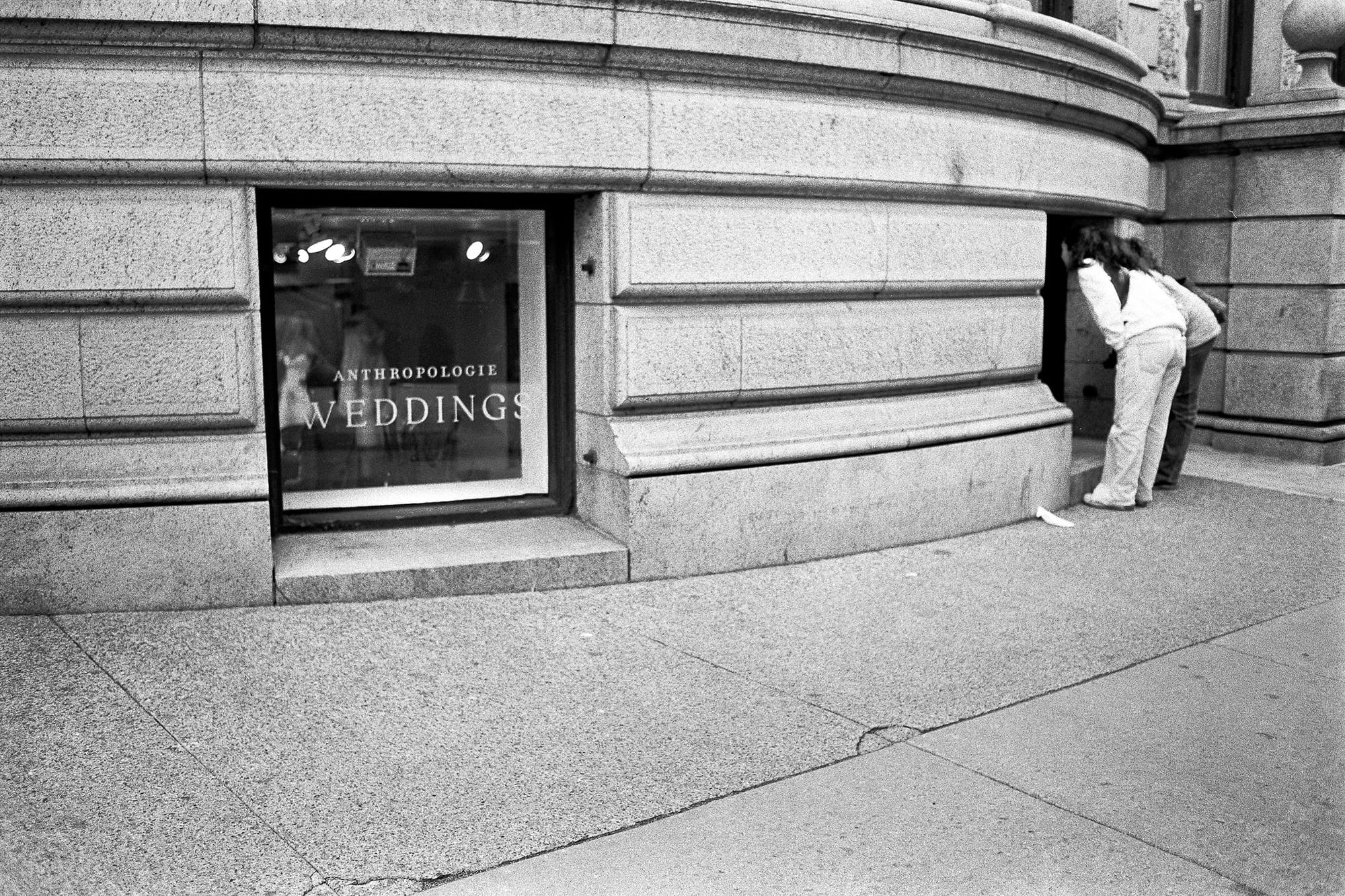 Two people bend to peer into a recess beside a stone shopfront window lettered Anthropologie Weddings