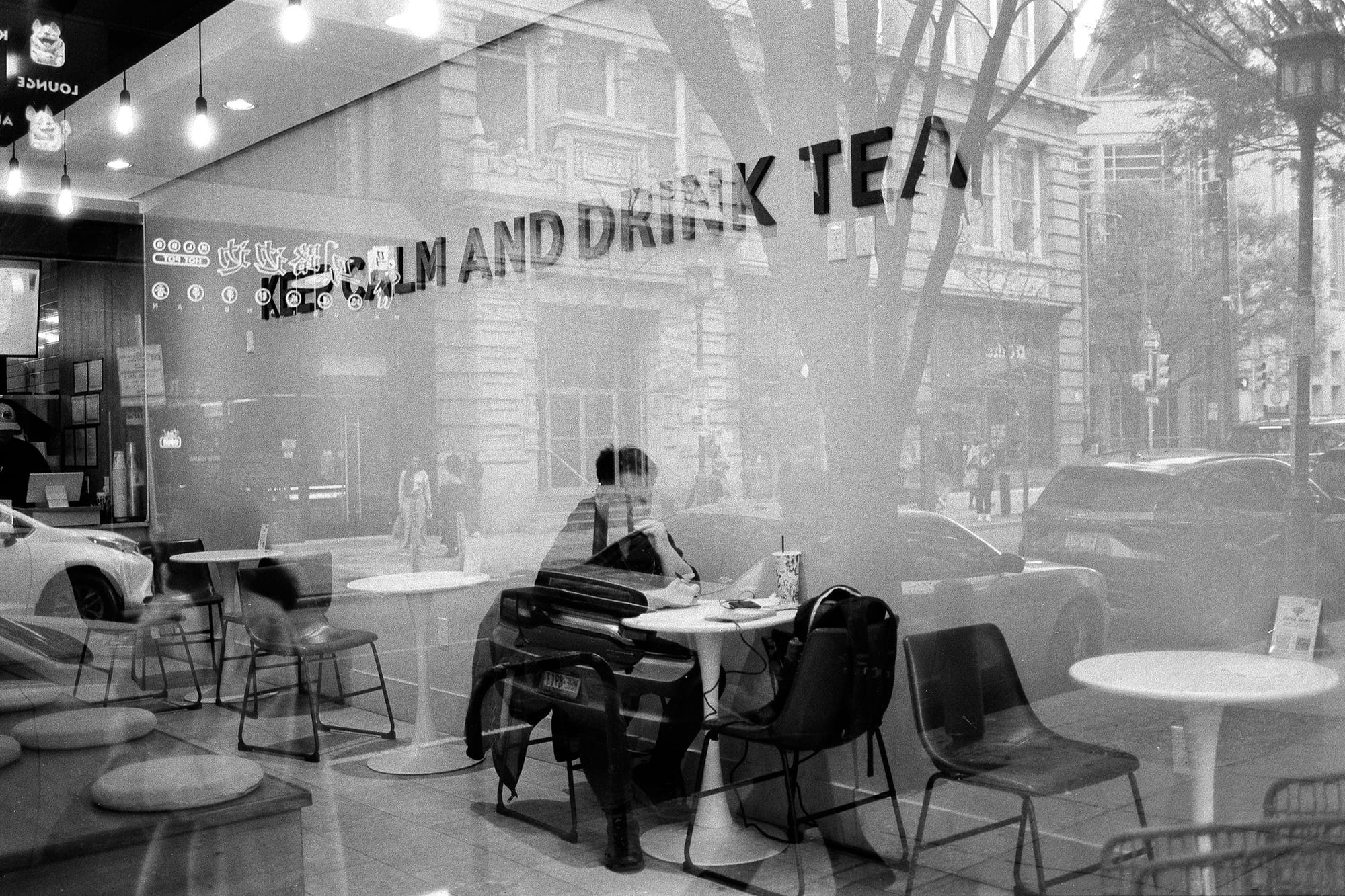 Seen through a café window lettered Keep Calm and Drink Tea, a person sits alone at a table with a drink as the street reflects across the glass