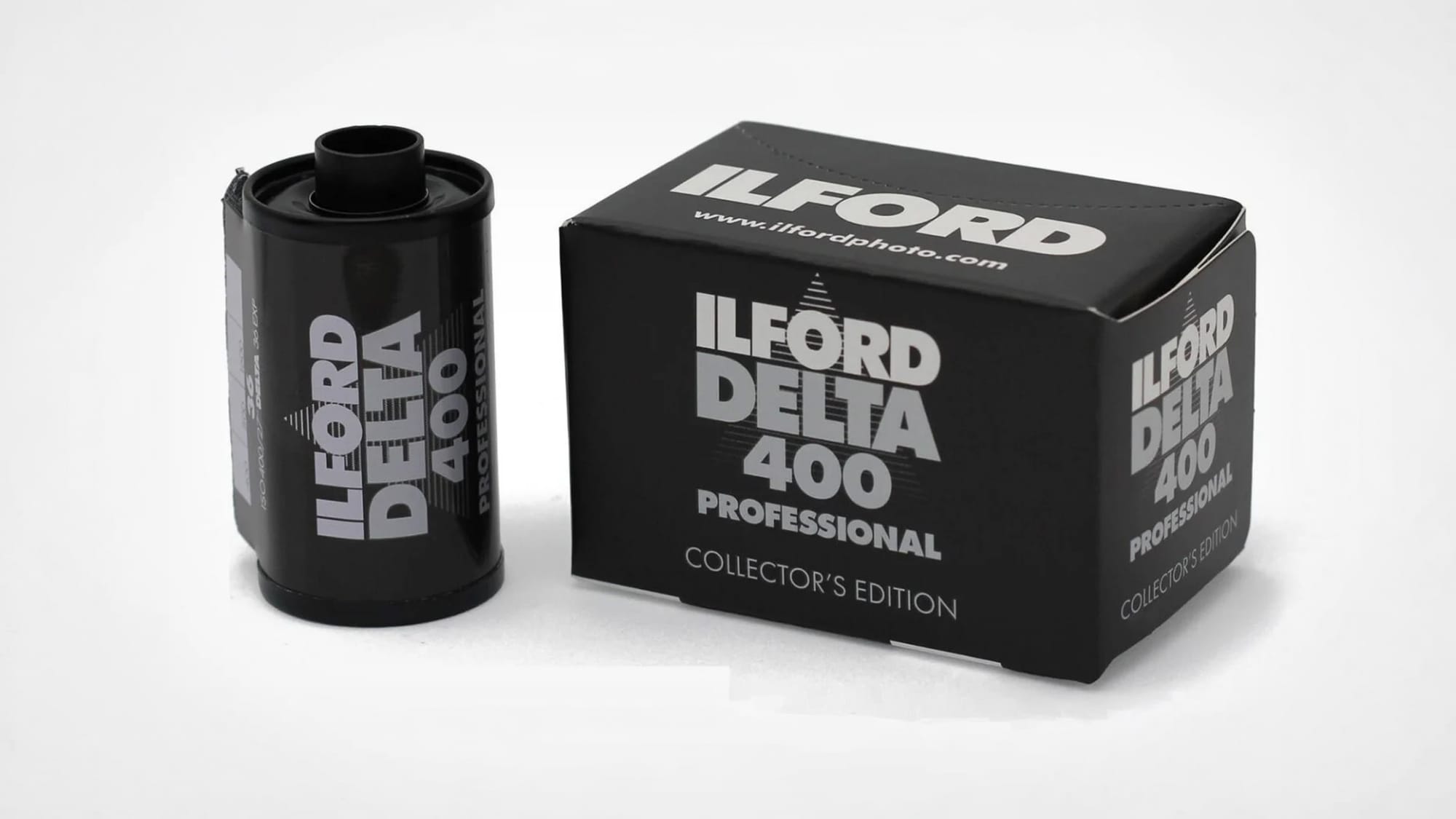 An Ilford Delta 400 Professional 135 cassette standing upright on a gray surface, in the standard white livery with black and green Delta lettering, marked 36 exposures and made in England.