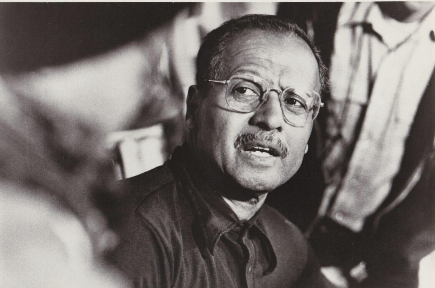 Roy DeCarava in close-up, wearing wire-rimmed glasses and a dark shirt, looking up and to his right in mid-sentence.